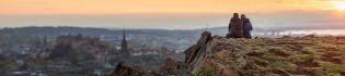 Two students sitting on a ledge of the Crags watching the sunset over the Edinburgh skyline.