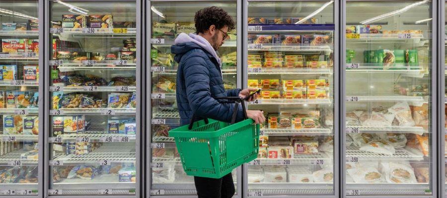Man with shopping basket standing next to frozen food freezer at store.