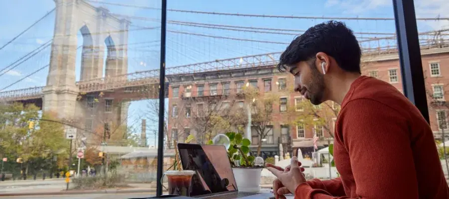 Student on a laptop in a cafe. Out the window in the background is the Brooklyn Bridge.