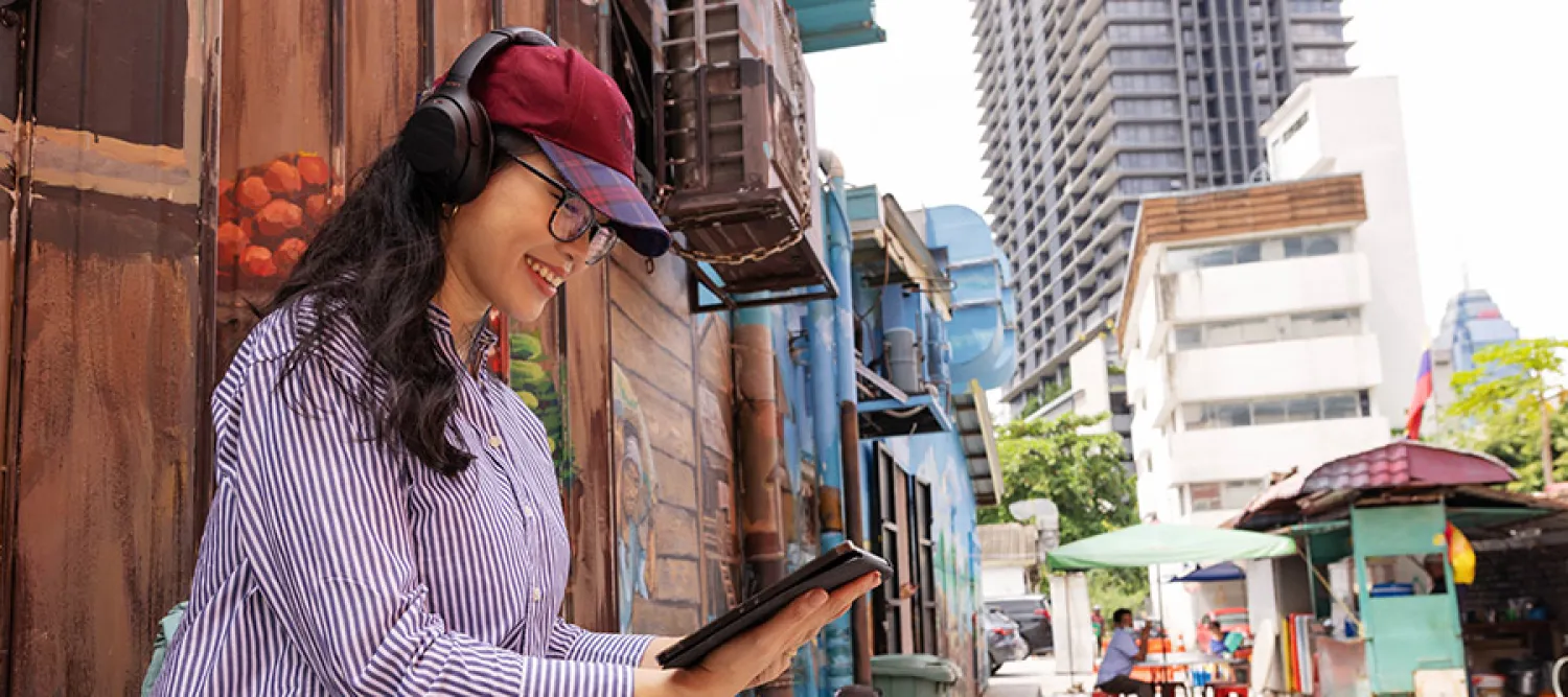 A student outside in a city holding a tablet and watching it with headphones.