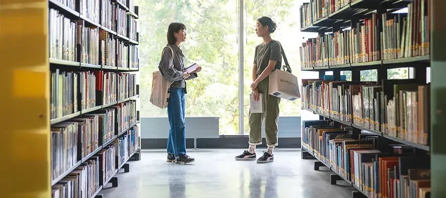Two students chatting at the library in between book shelves.