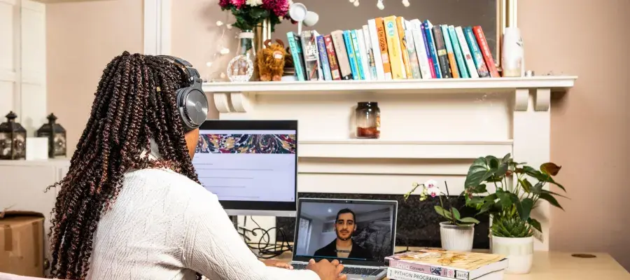 Woman sits at desk wearing headphones and looking at a monitor and a laptop, with stack of computer programming books beside her.