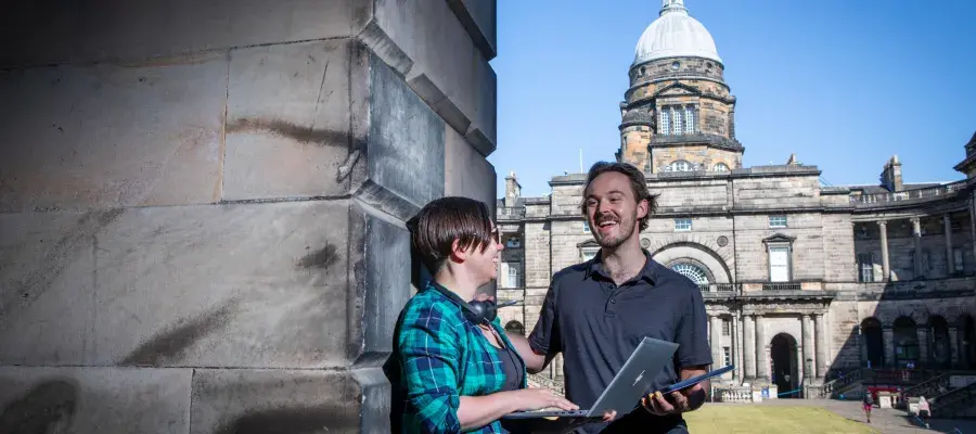 One male and one female student stand outside Old College talking and using a laptop.