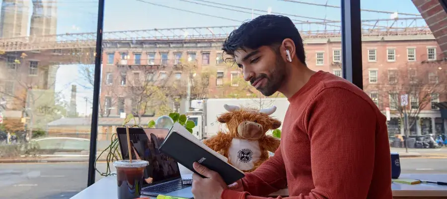 Young man sits in cafe wearing headphones and reading a notebook, with a laptop, coffee and University of Edinburgh soft toy.