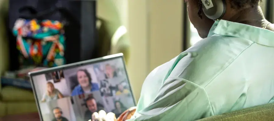 Woman sits in chair wearing headphones using a laptop, while on a video call with a group of people.