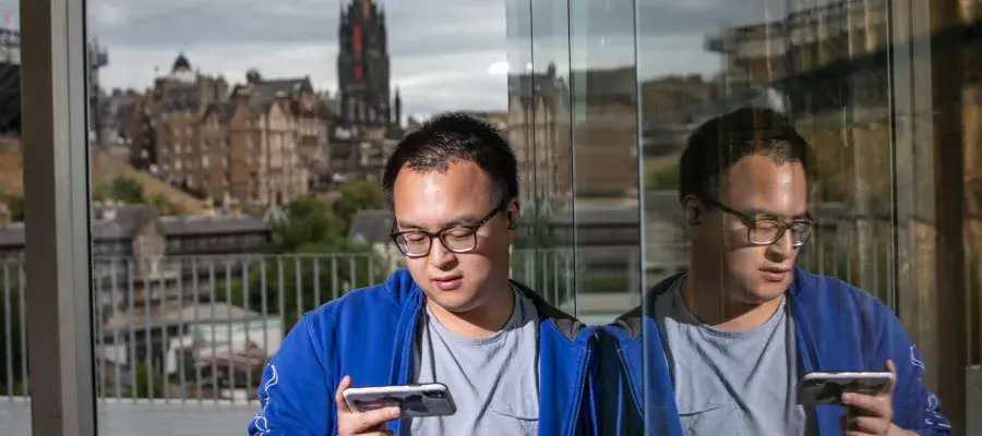 Man stands outside building with Edinburgh cityscape in the background, looking at a mobile phone.
