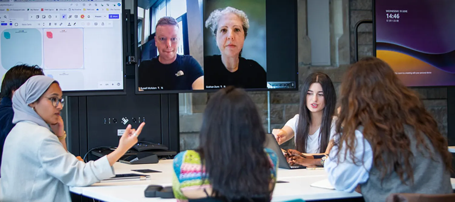 Students sit at a table in front of a large screen showing a Teams call.