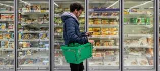 Man with shopping basket standing next to frozen food freezer at store.