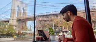 Student on a laptop in a cafe. Out the window in the background is the Brooklyn Bridge.
