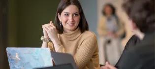 A woman smiling at a another person while sitting in front of a laptop.