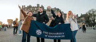 Four students holding a blue banner that says the University of Edinburgh in Front of Edinburgh Castle.