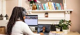 Woman sits at desk wearing headphones and looking at a monitor and a laptop, with stack of computer programming books beside her.