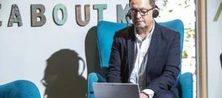 Middle-aged man in suit and glasses sits in chair wearing headphones and looking at a tablet.