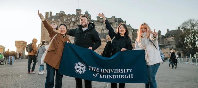 Four students holding a blue banner that says the University of Edinburgh in Front of Edinburgh Castle.
