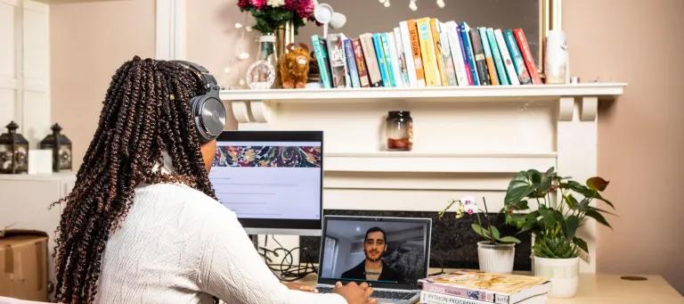 Woman sits at desk wearing headphones and looking at a monitor and a laptop, with stack of computer programming books beside her.