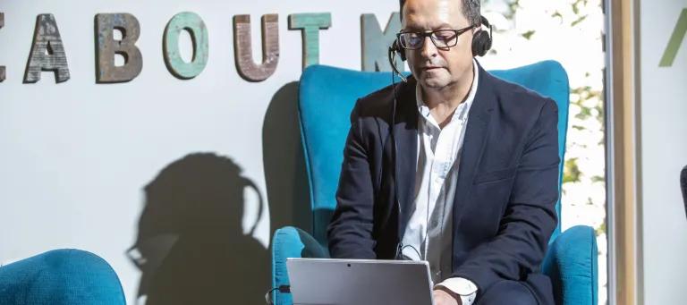 Middle-aged man in suit and glasses sits in chair wearing headphones and looking at a tablet.