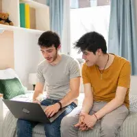 Two students sit on a bed looking at a laptop in their university accommodation.