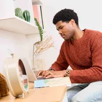A photo of a male student sitting at a desk, in his room, typing on his computer at Buccleuch Place accommodation