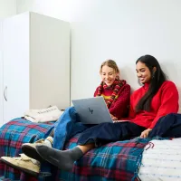 A photo of two women sitting on a university accommodation bed. They are both smiling while looking at laptop, which is resting on top of the legs of one of the two women.