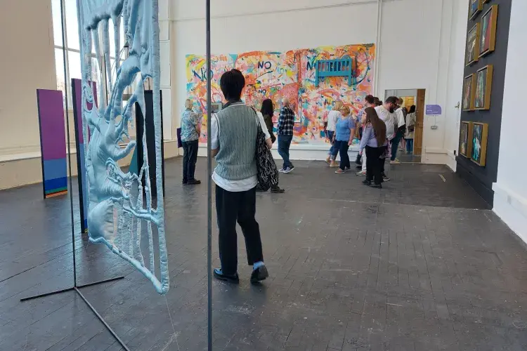 Exhibition shot showing blue fabric handing over a metal frame in the foreground to the right. A male presenting person walks towards a group of people in the background.