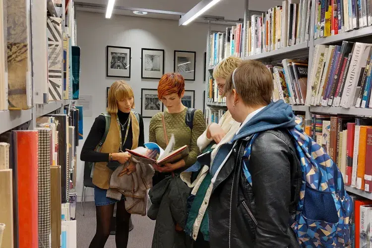 A group of people look at a book in the ECA library. They are surrounded by shelves filled with colourful books.