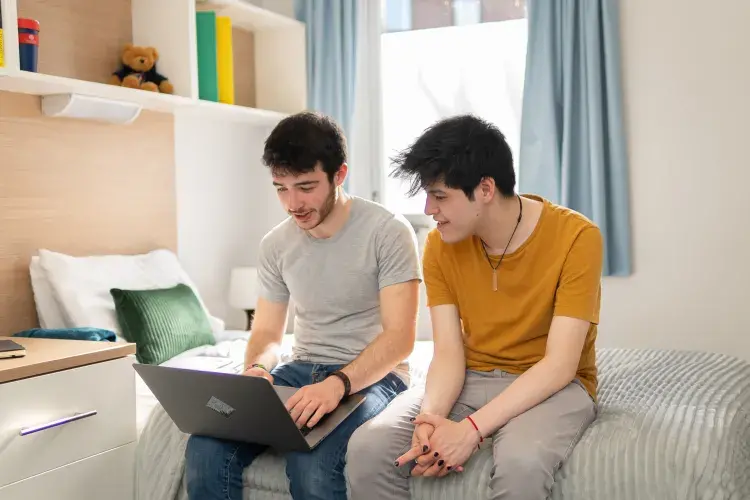 Two students sit on a bed looking at a laptop in their university accommodation.
