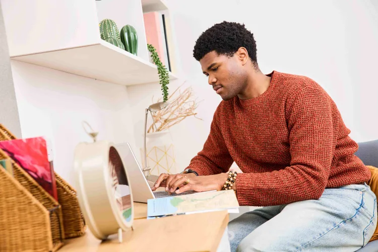 A photo of a male student sitting at a desk, in his room, typing on his computer at Buccleuch Place accommodation