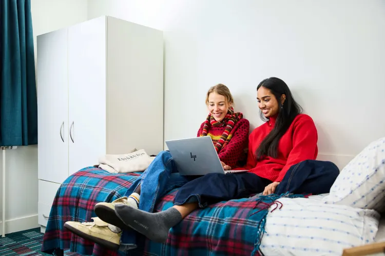 A photo of two women sitting on a university accommodation bed. They are both smiling while looking at laptop, which is resting on top of the legs of one of the two women.