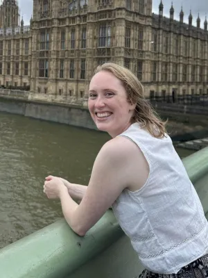 Photo shows Kimberly standing on a bridge in front of the Houses of Parliament, smiling at the camera and wearing a white shirt.