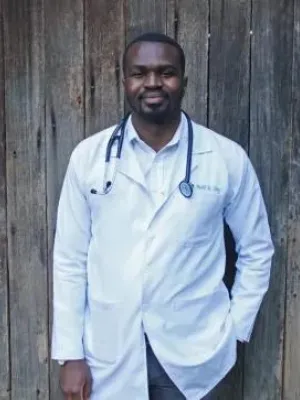 A doctor with a stethoscope round his neck, with a lab coat on, standing against a brown fence.