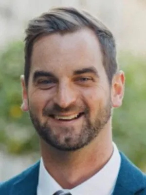 Headshot of man smiling at camera, wearing a suit against a natural back drop